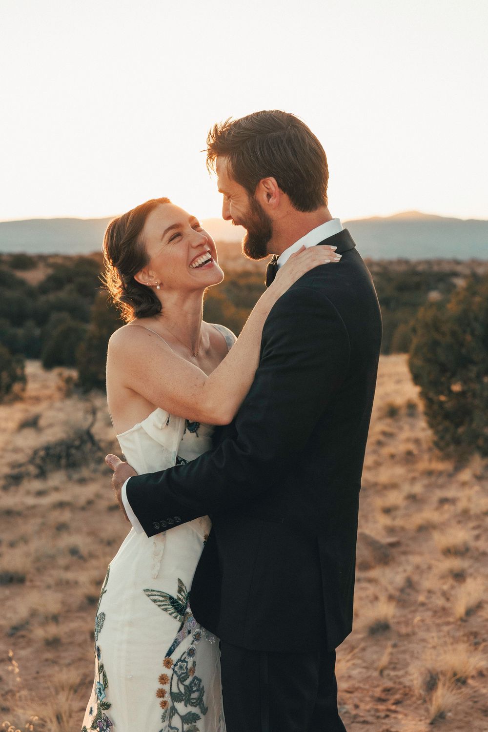 Laughing bride in floral wedding gown embraces groom in black tuxedo at desert sunset.