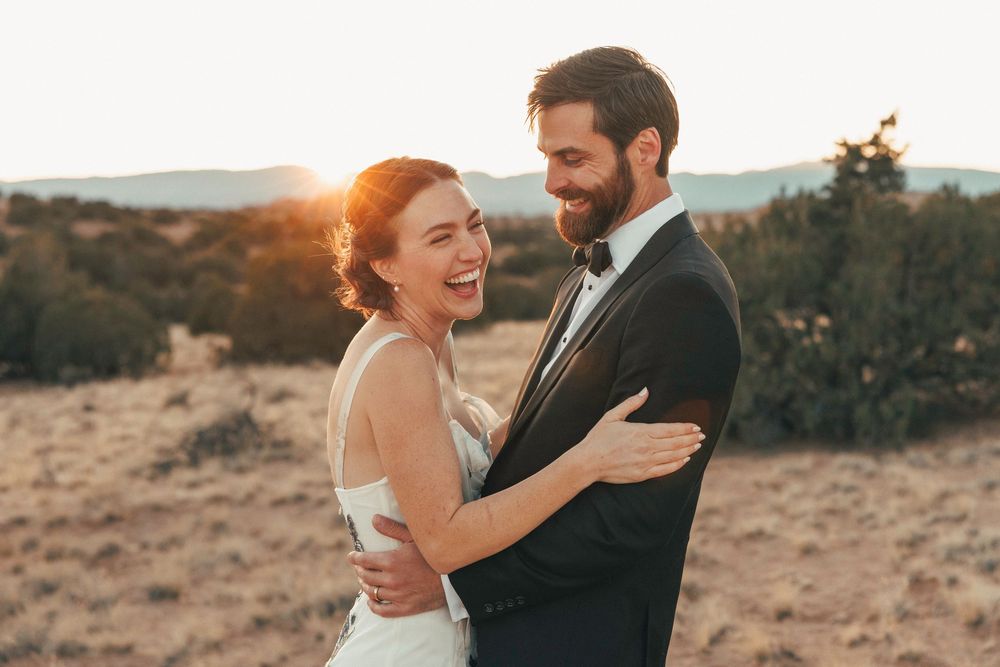 Laughing bride and smiling groom embrace in a sun-drenched desert field at golden hour.