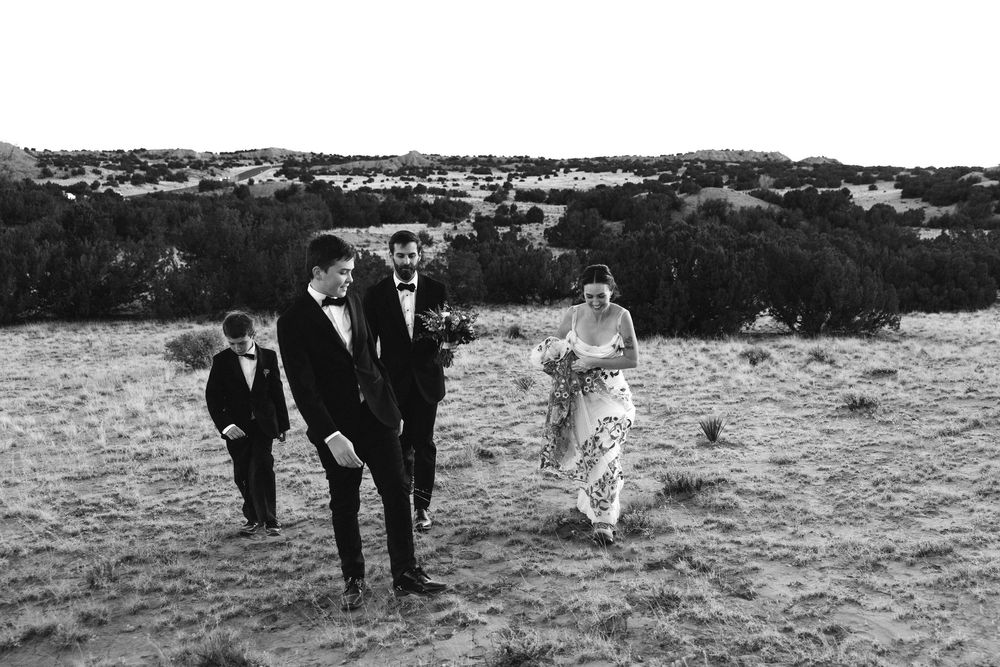 Black and white photo of a wedding party walking through a field with trees in the background.