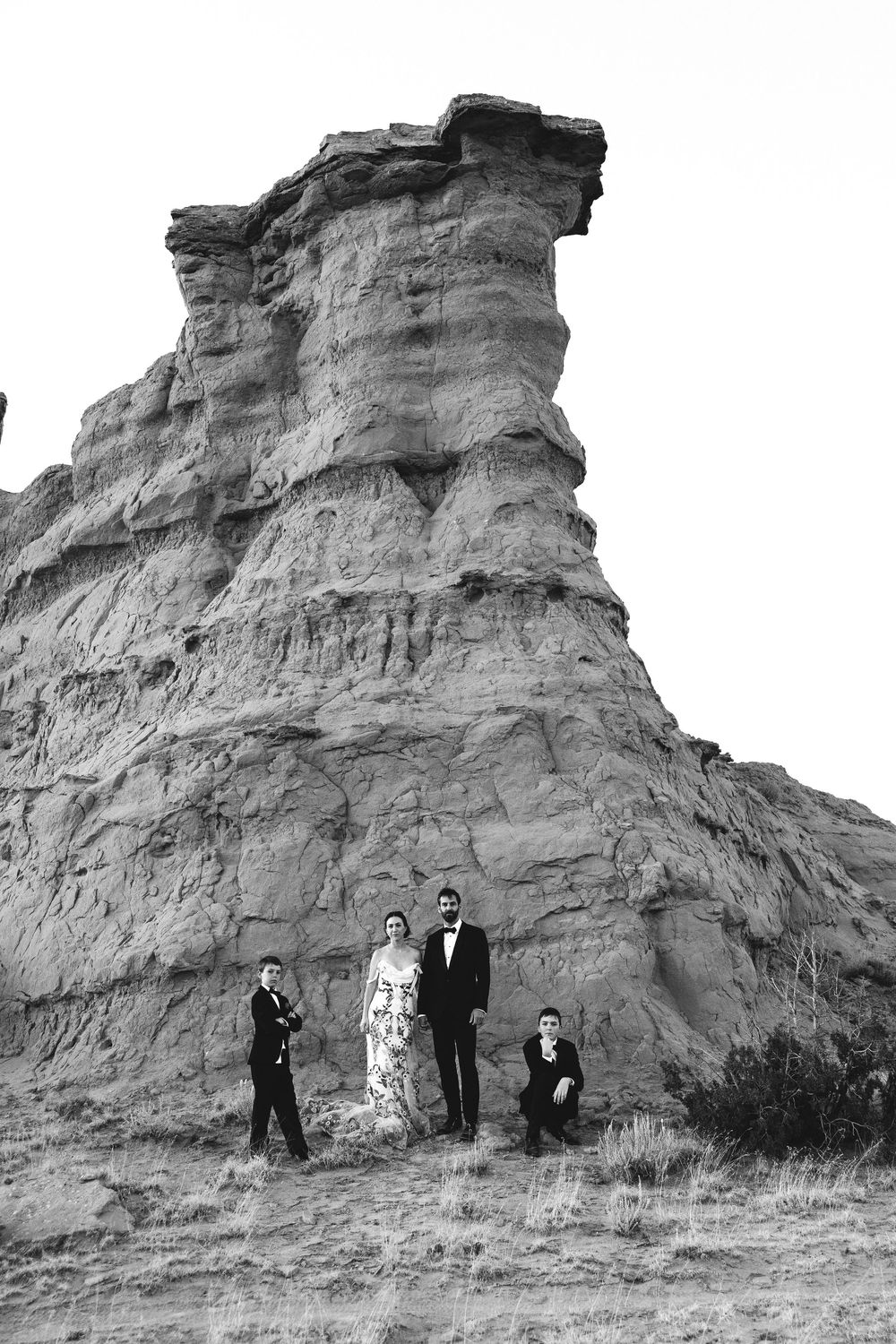 Black and white photo of a wedding party posing beneath a tall sandstone rock formation.