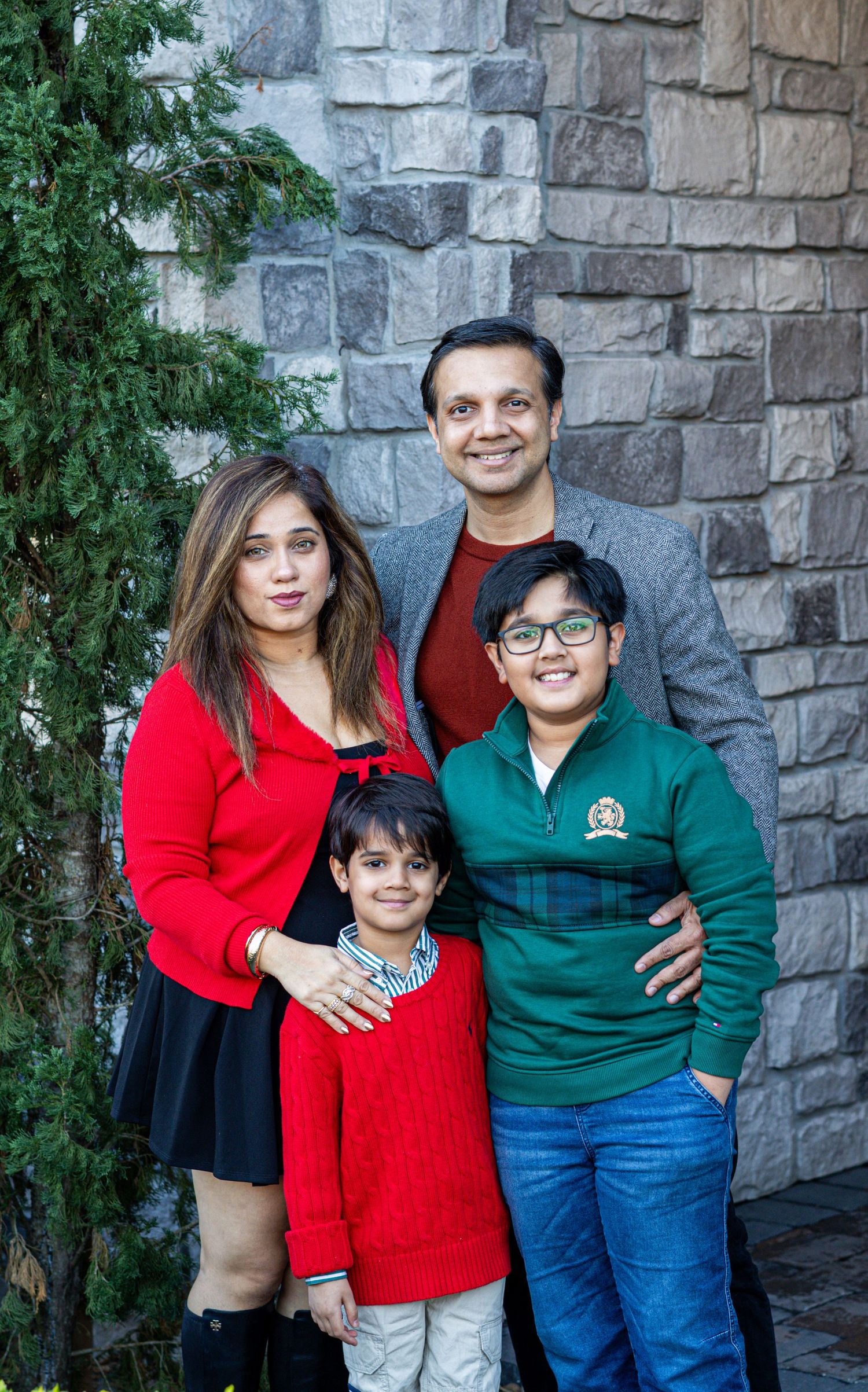 Family of four posing together in front of a stone wall with greenery, wearing colorful sweaters.