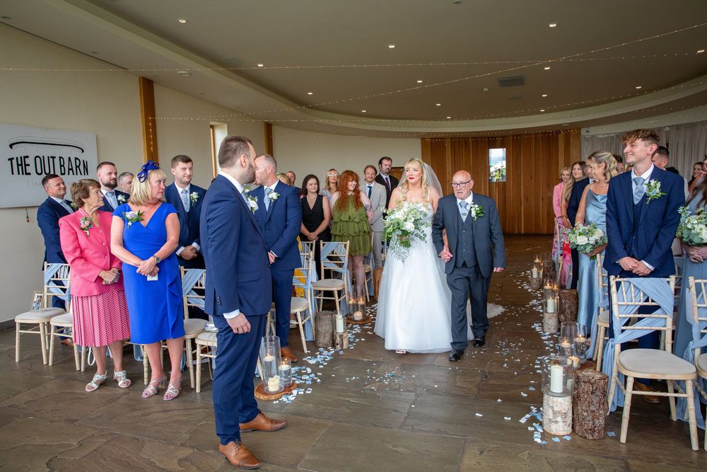 Bride walks down the aisle toward groom in modern barn ceremony venue filled with seated guests.