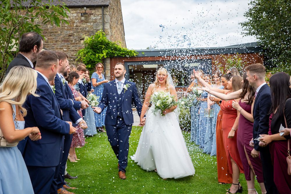 Bride and groom walk hand in hand through colorful confetti shower thrown by cheering wedding guests.