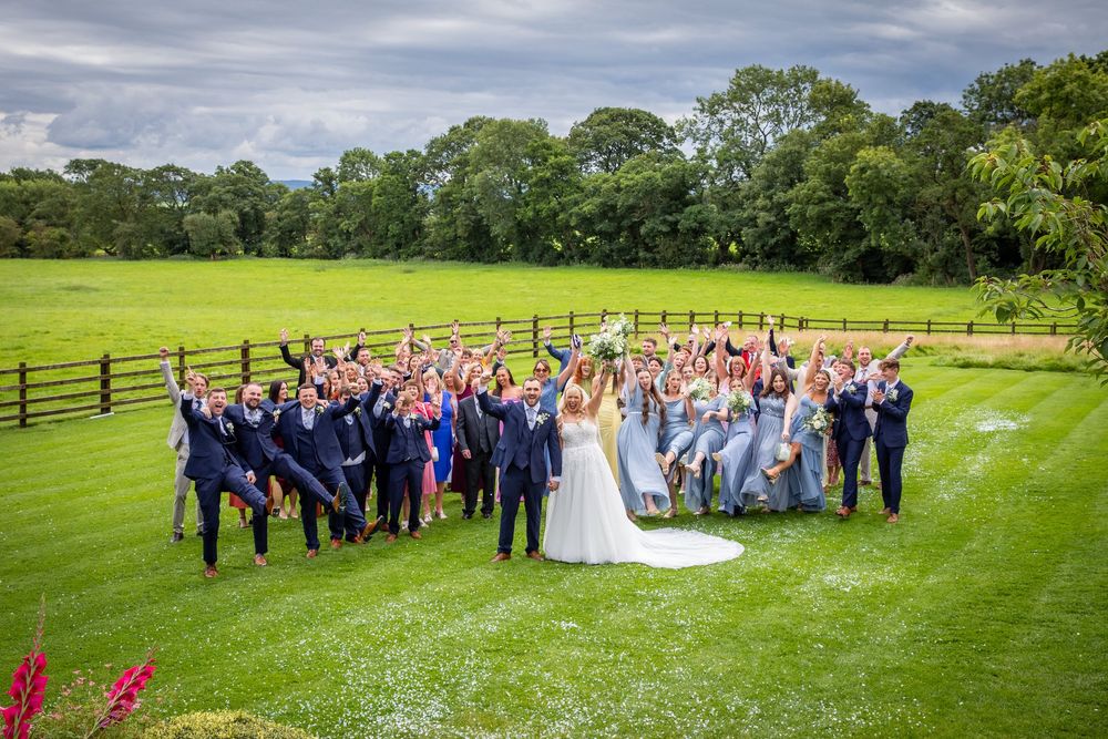 Large wedding party celebrating outdoors on lush green lawn with bride and groom centered among cheering guests.