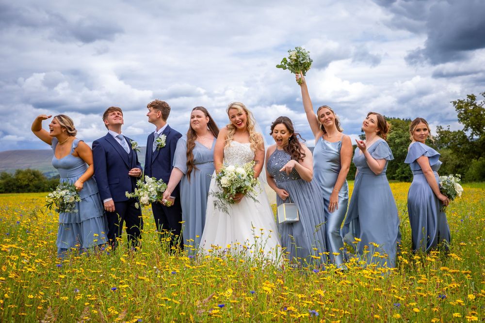 Bride with bridesmaids in blue dresses celebrating in a wildflower meadow under dramatic cloudy sky.