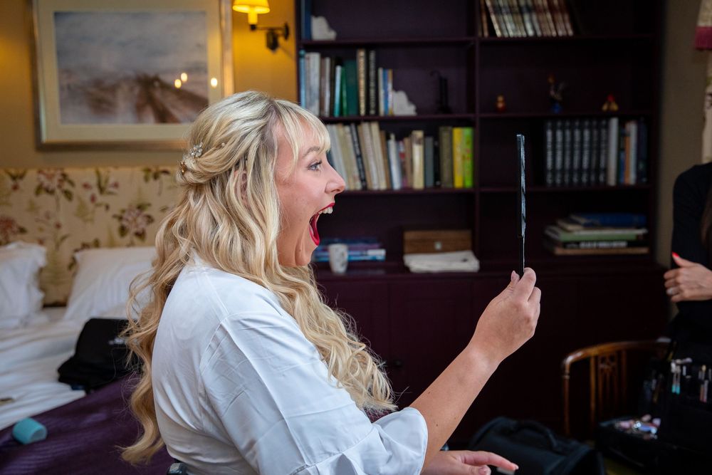 Excited blonde bride in white robe reacts with joy while looking at phone during wedding preparations.