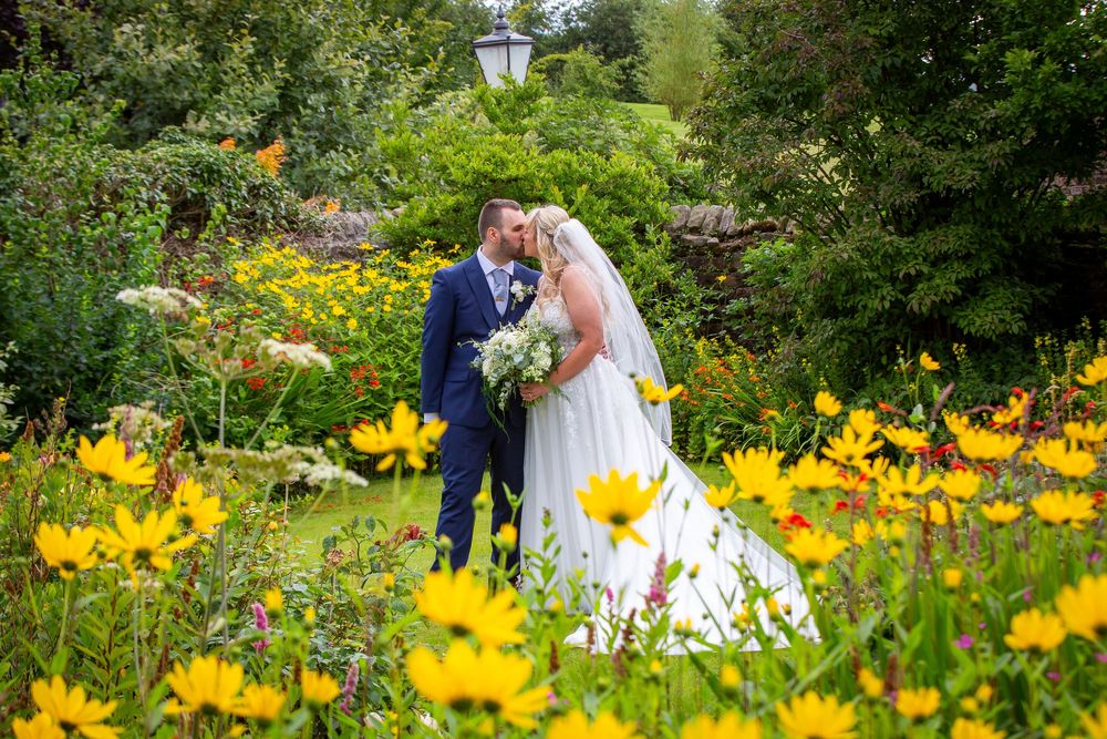 Bride and groom embracing romantically surrounded by vibrant yellow flowers in a lush garden setting.