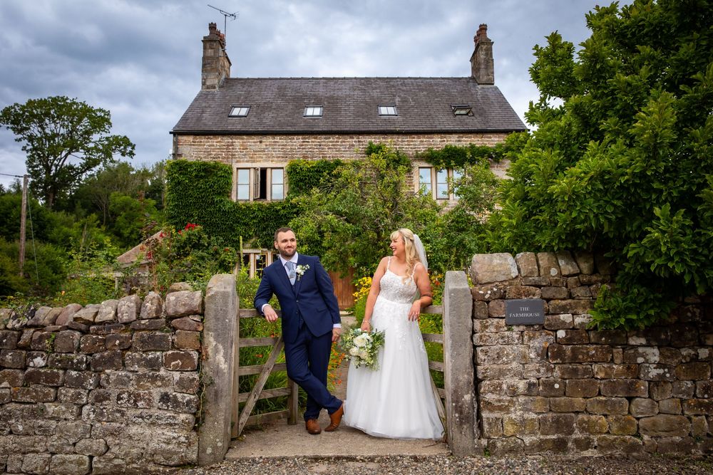 Bride and groom walking through rustic stone gate in front of charming ivy-covered country house.