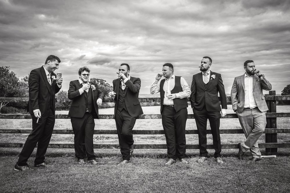 Black and white photo of groom and groomsmen posing casually against wooden fence in countryside.