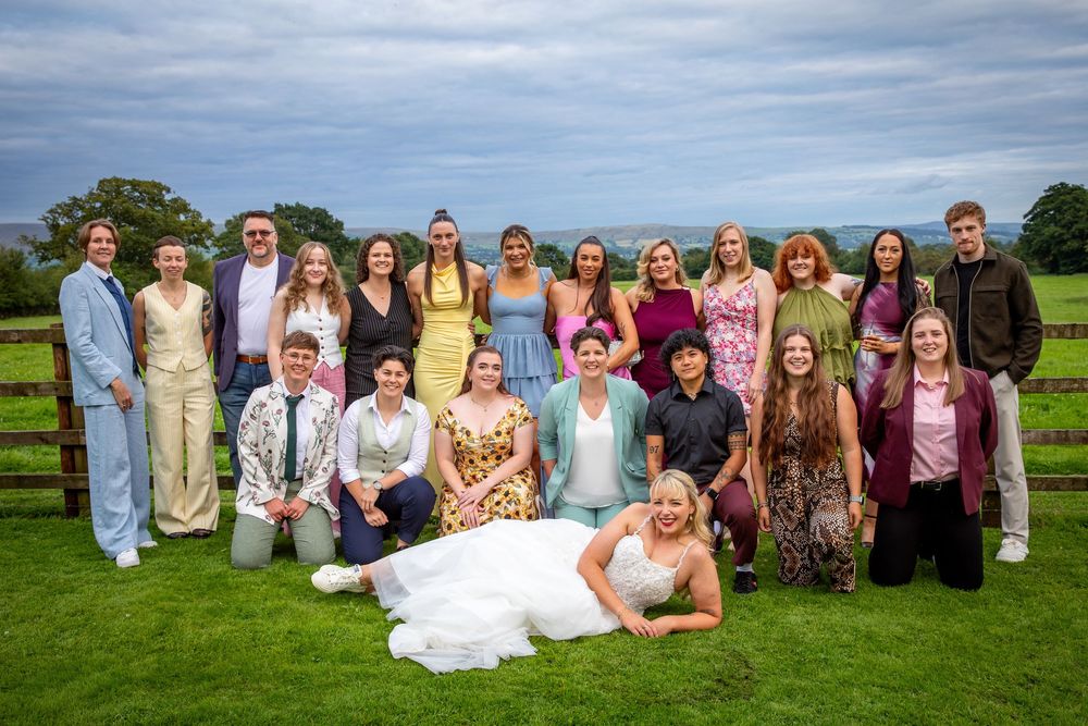Bride posing with large group of wedding guests on green lawn with scenic countryside backdrop.