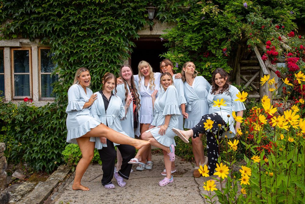 Bridal party in matching grey robes pose playfully outside ivy-covered venue with bright yellow flowers.
