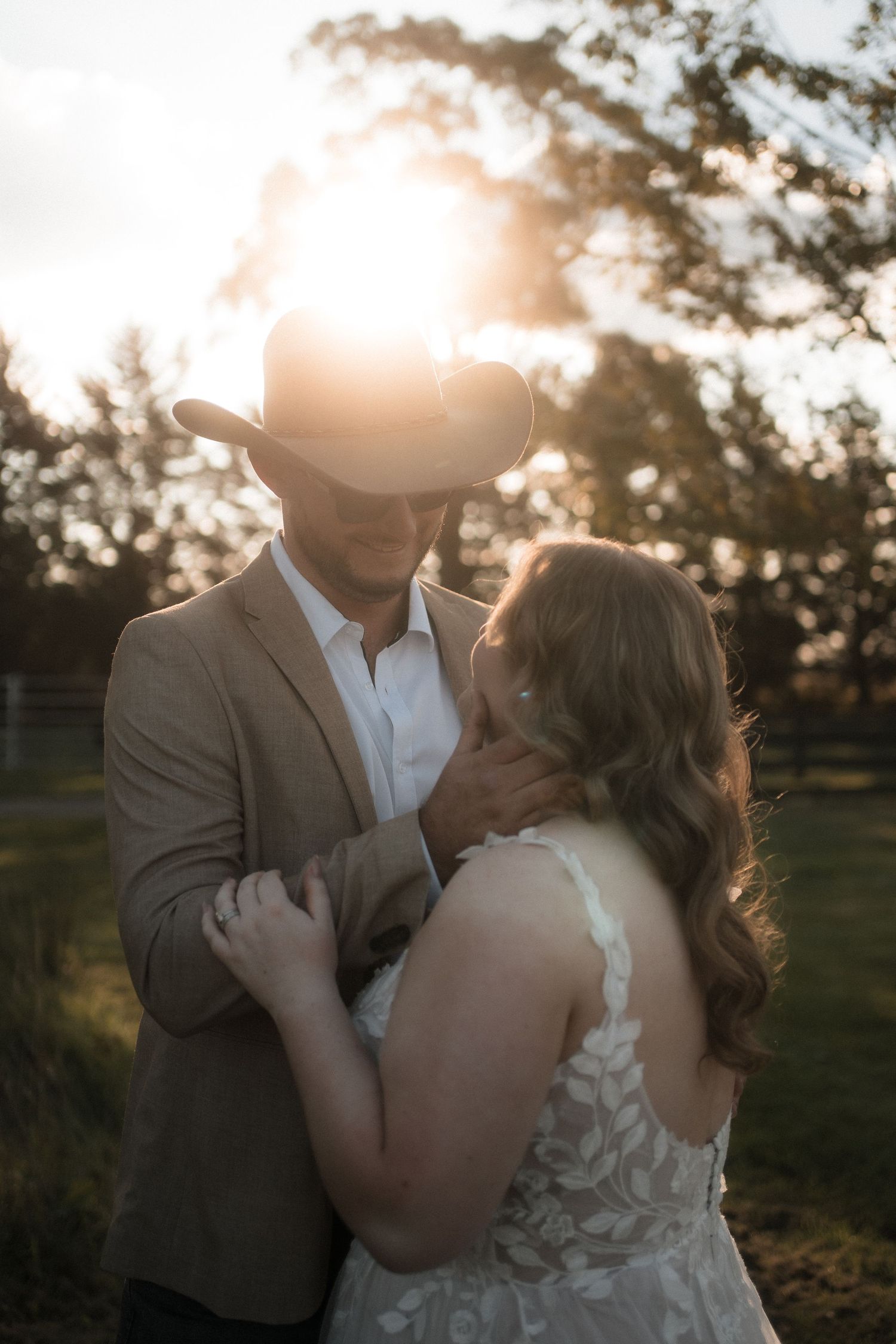 A romantic sunset photo of a couple in wedding attire embracing in a dreamy outdoor setting with golden light filtering through trees.