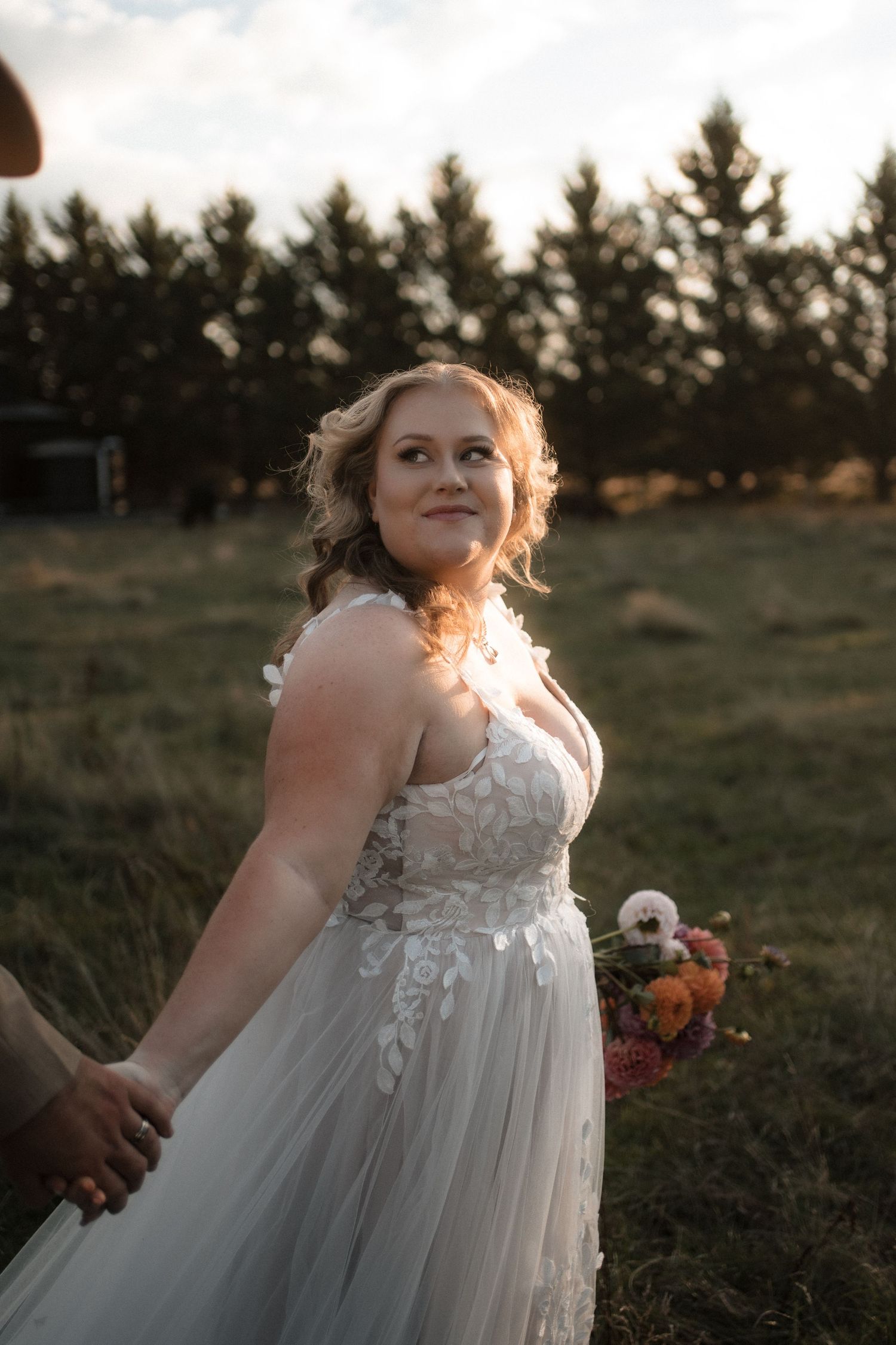 A bride in a flowing white lace dress twirls joyfully in a golden-lit field at sunset.