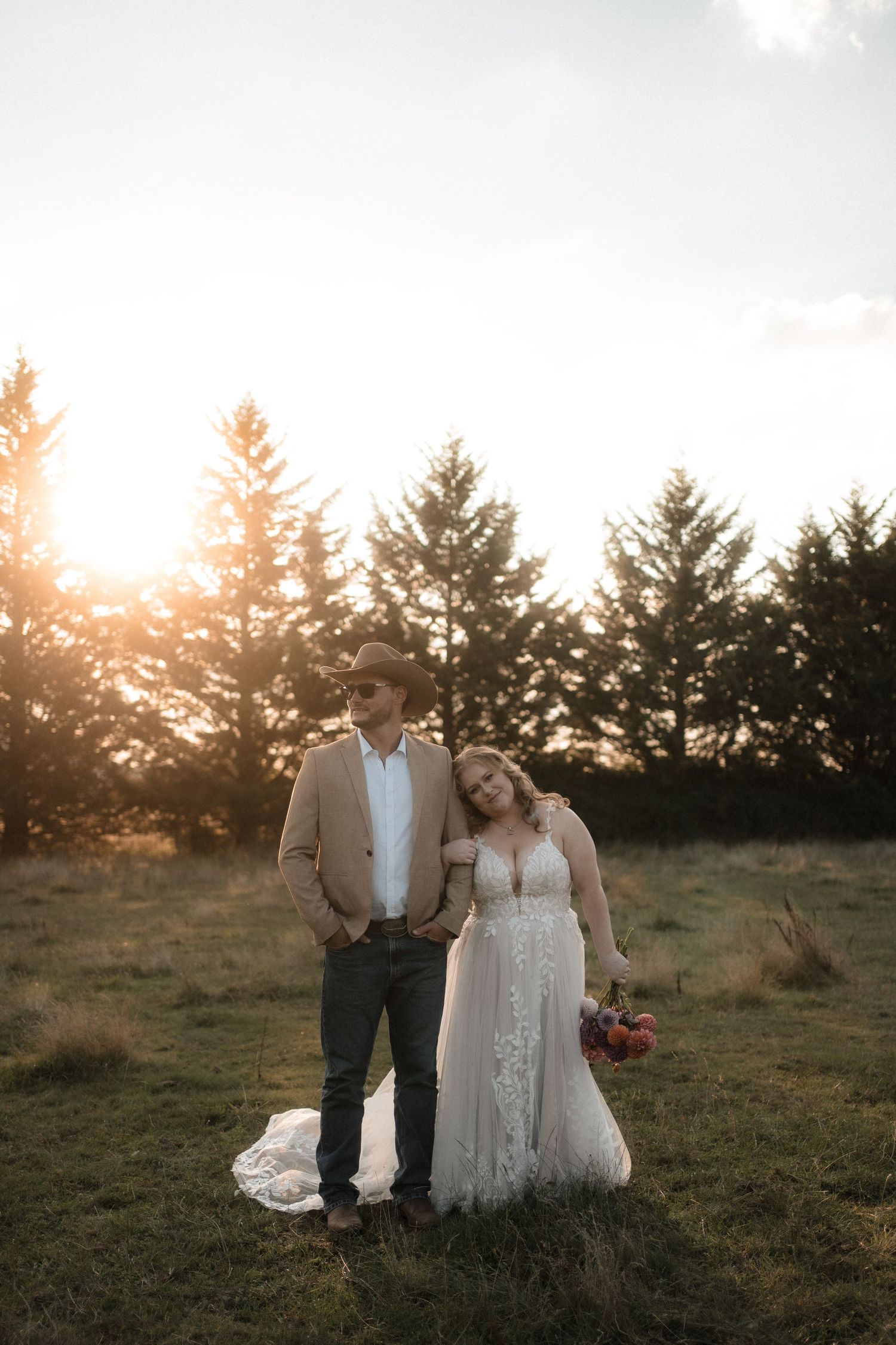A bride and groom share a romantic moment at sunset in a field with evergreen trees in the background.