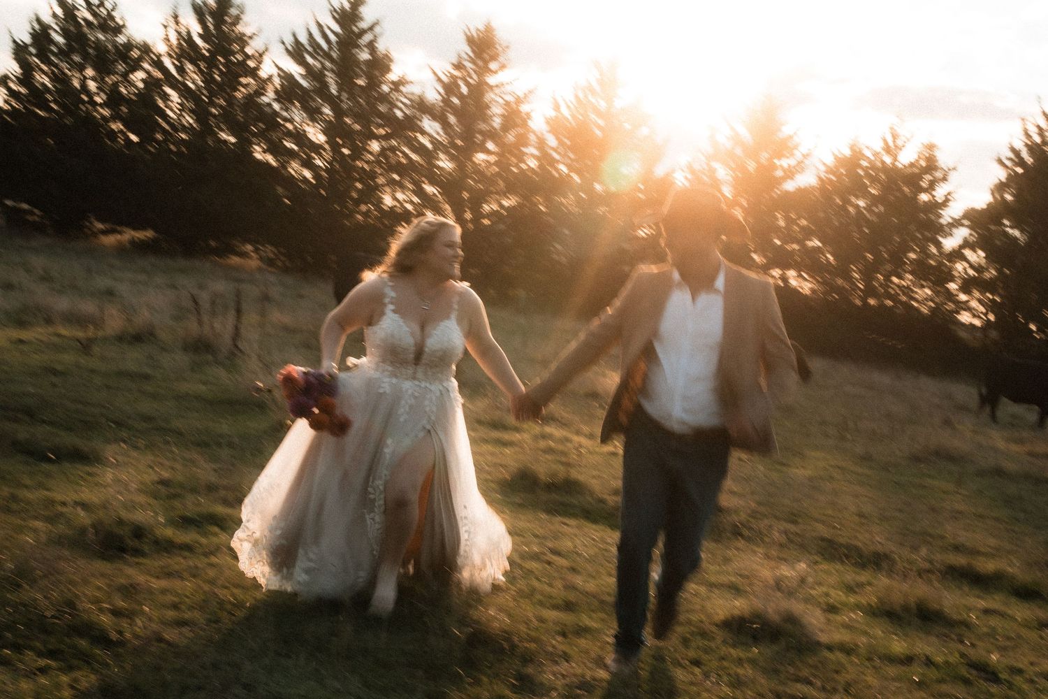 A couple walks hand in hand through a golden sunset field as their wedding dress and attire flow in the breeze.