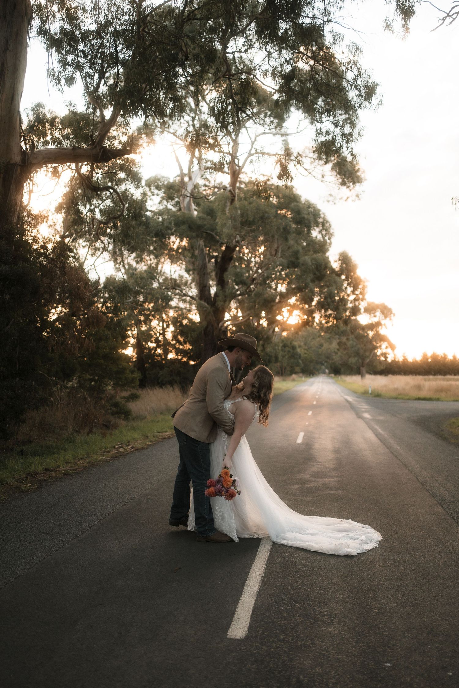 A romantic sunset silhouette of a couple in wedding attire embracing on a tree-lined country road.