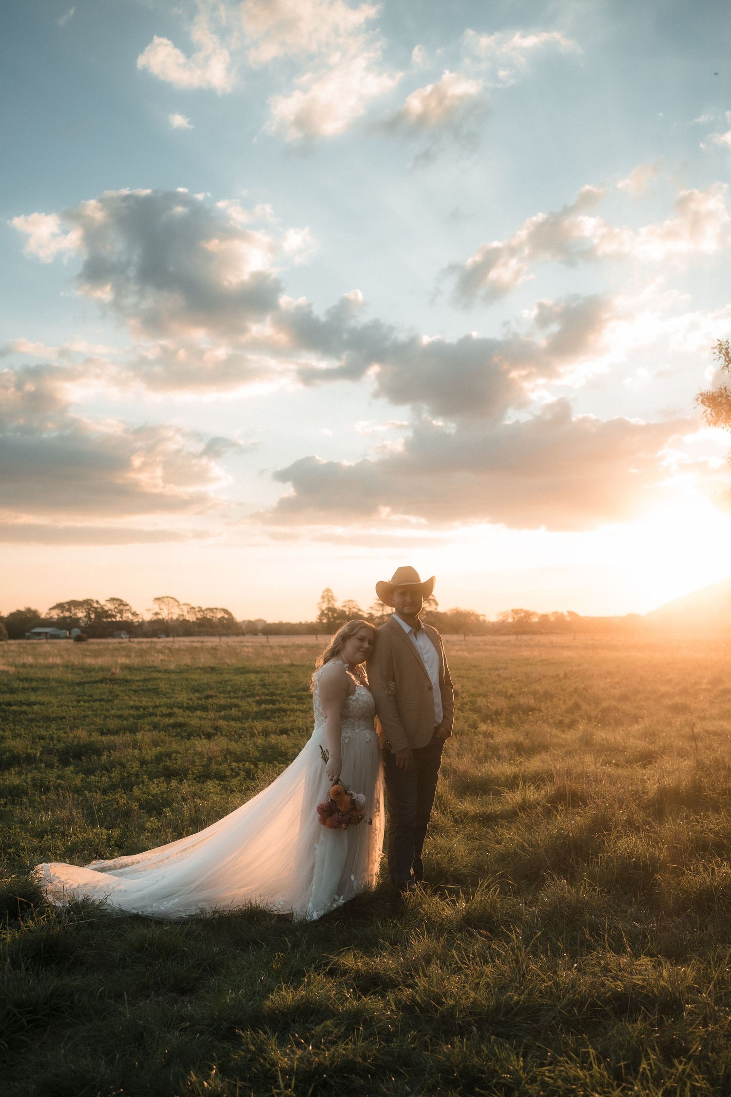 A couple in wedding attire stands in a golden field at sunset with dramatic clouds in the background.