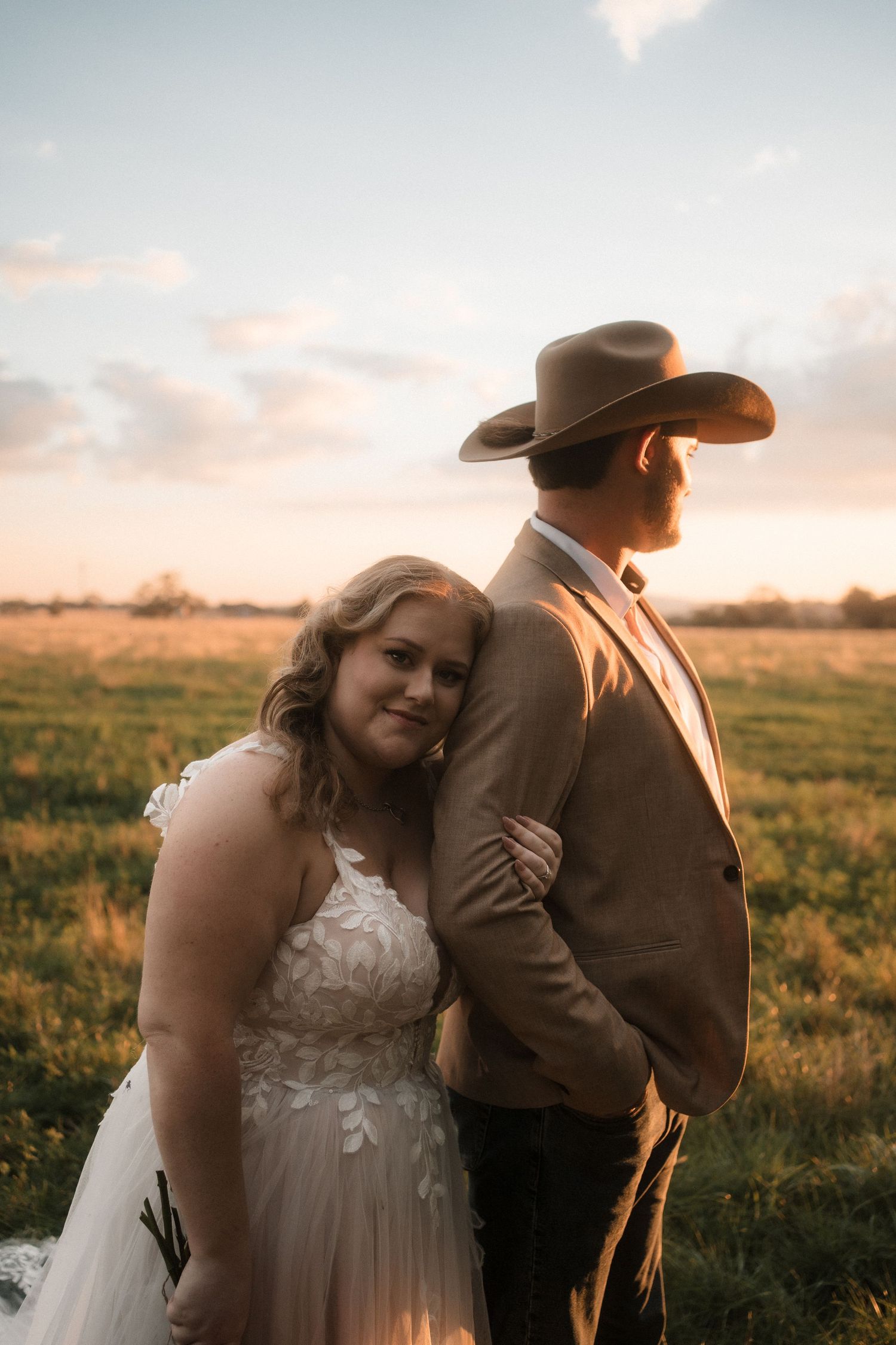 A couple shares an intimate moment in a field at sunset wearing western attire and wedding clothes.