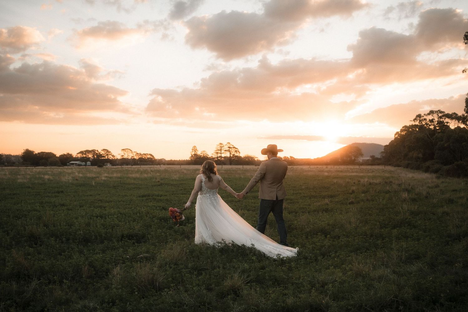 A romantic wedding couple stands in a golden field at sunset while their white dress flows in the breeze.