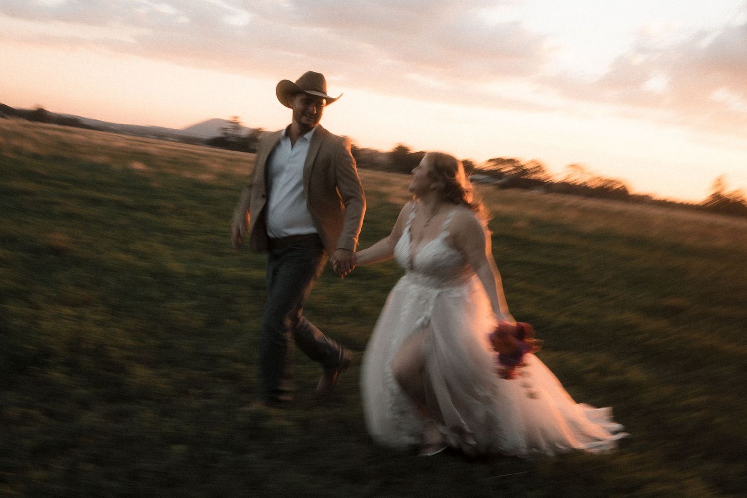 Couple walks hand in hand through a scenic meadow at golden sunset with mountains in the background.
