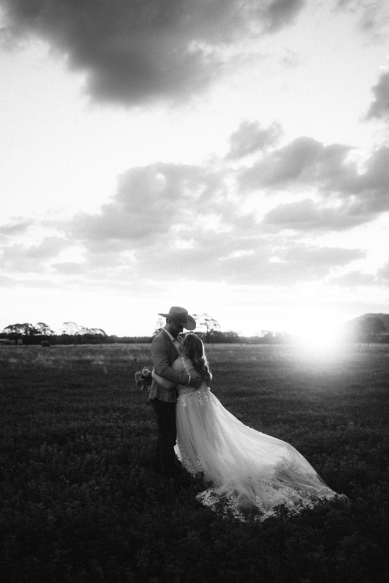 A couple in wedding attire embraces in a dramatic black and white landscape at sunset with moody clouds overhead.