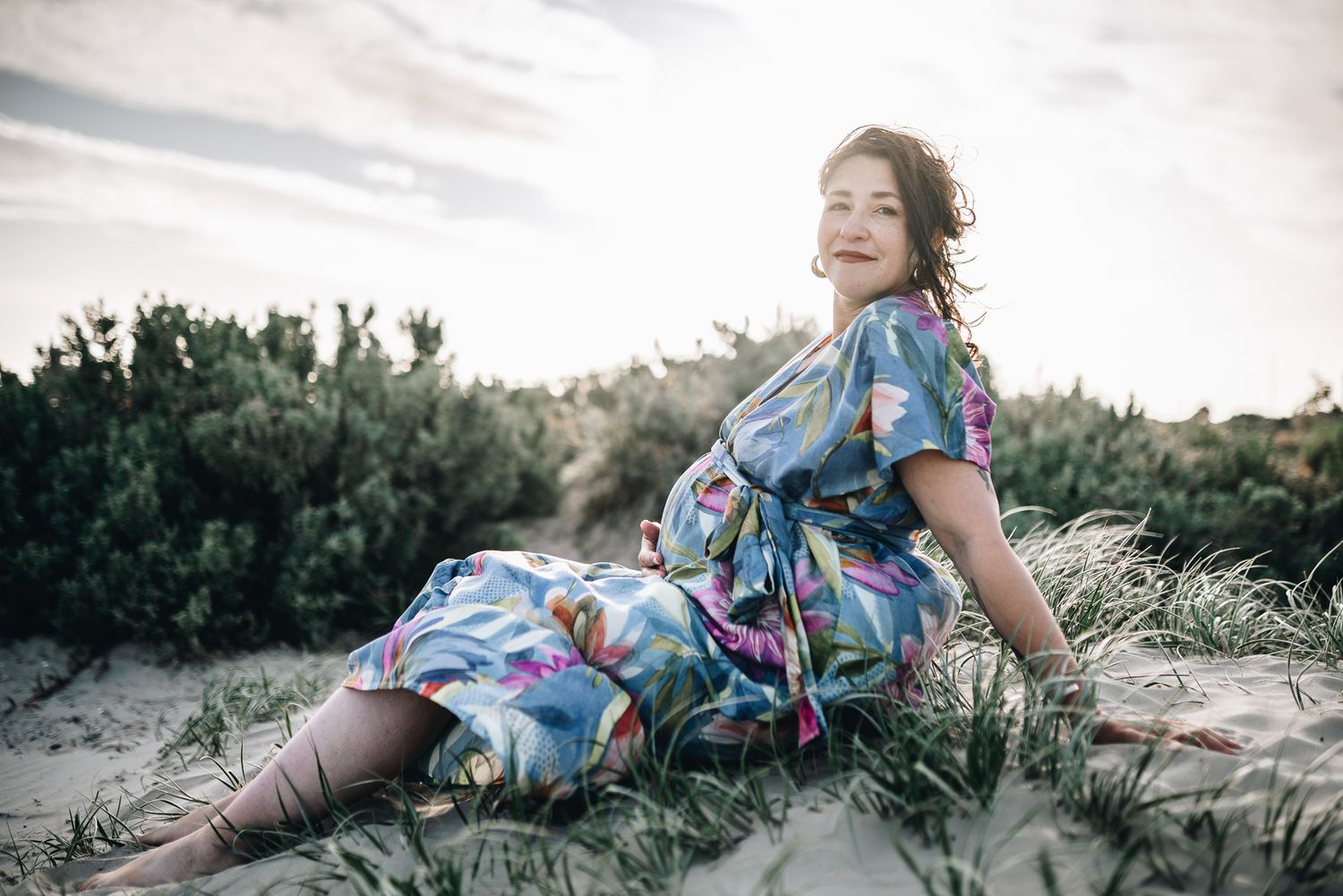 A pregnant lady in a colorful floral dress sits on a grassy sand dune during a Melbourne maternity photo session.