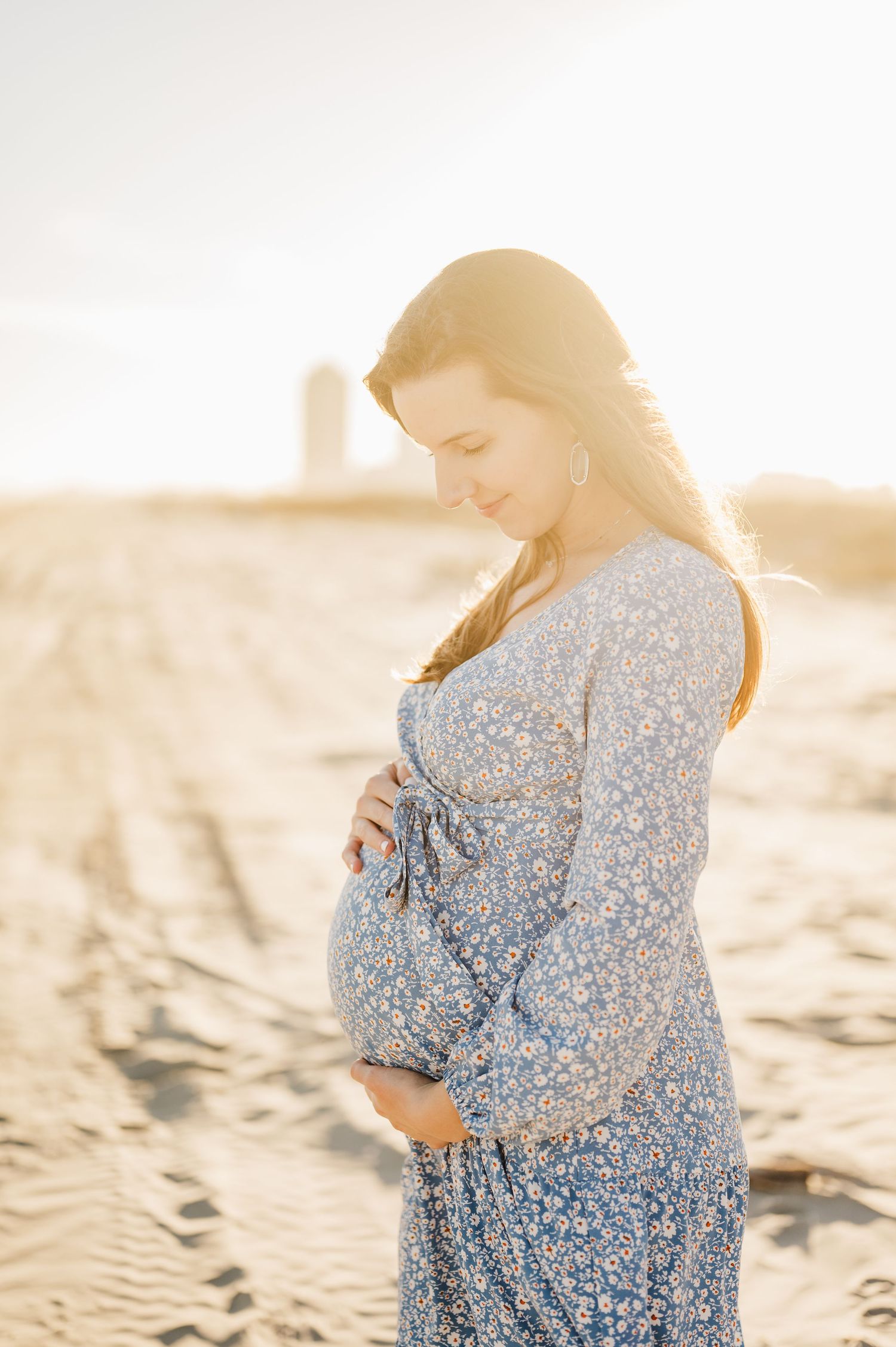 Expectant mother in sparkly blue dress poses on beach at sunset with golden light illuminating the scene.