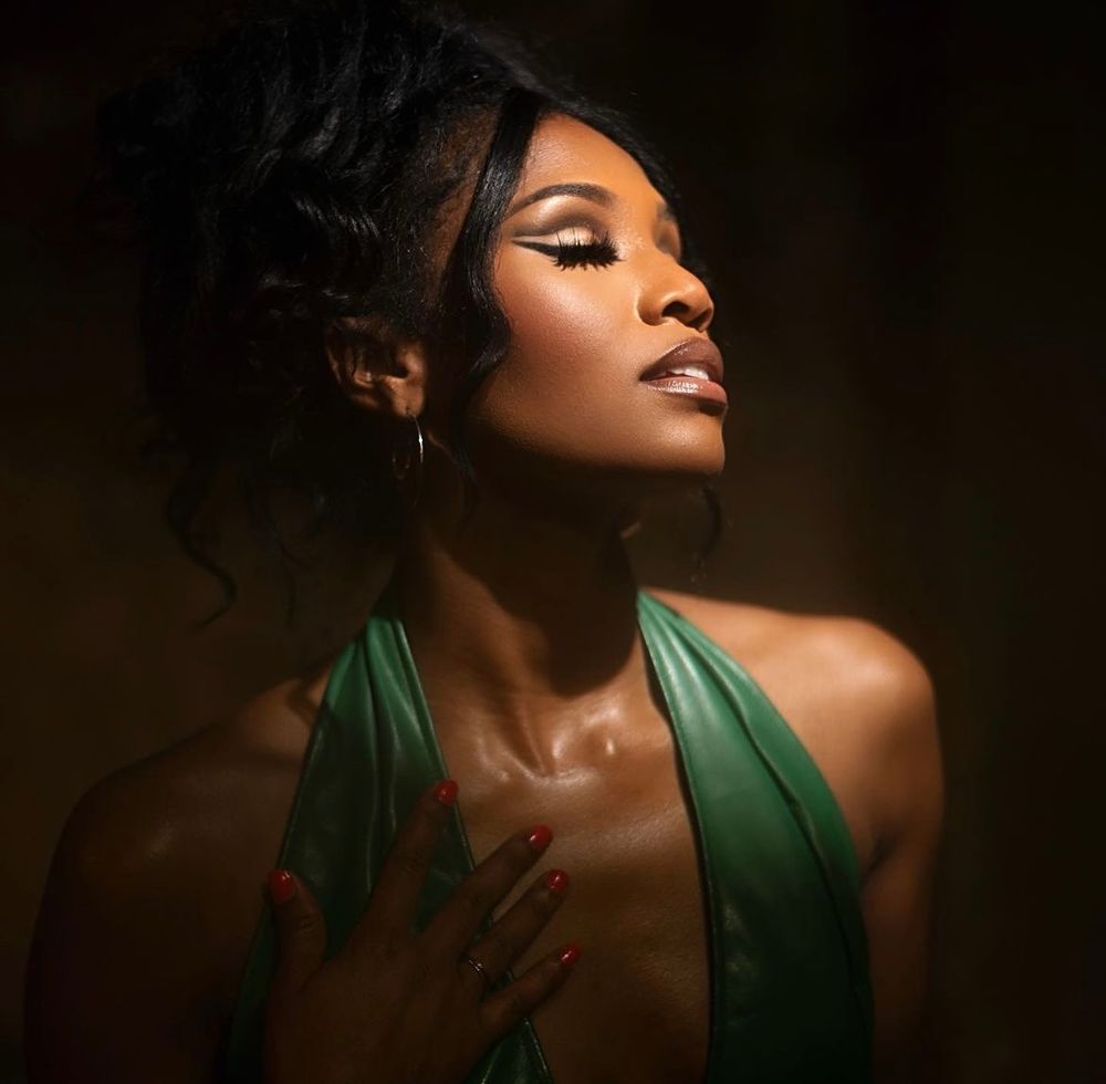 A Black woman in a green halter dress poses gracefully with eyes closed, hand on chest, in dramatic studio lighting.