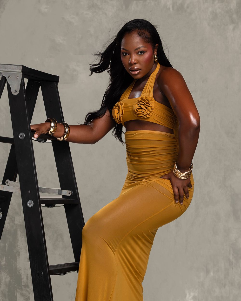 Woman in mustard yellow crop top and maxi skirt posing confidently beside a black ladder.