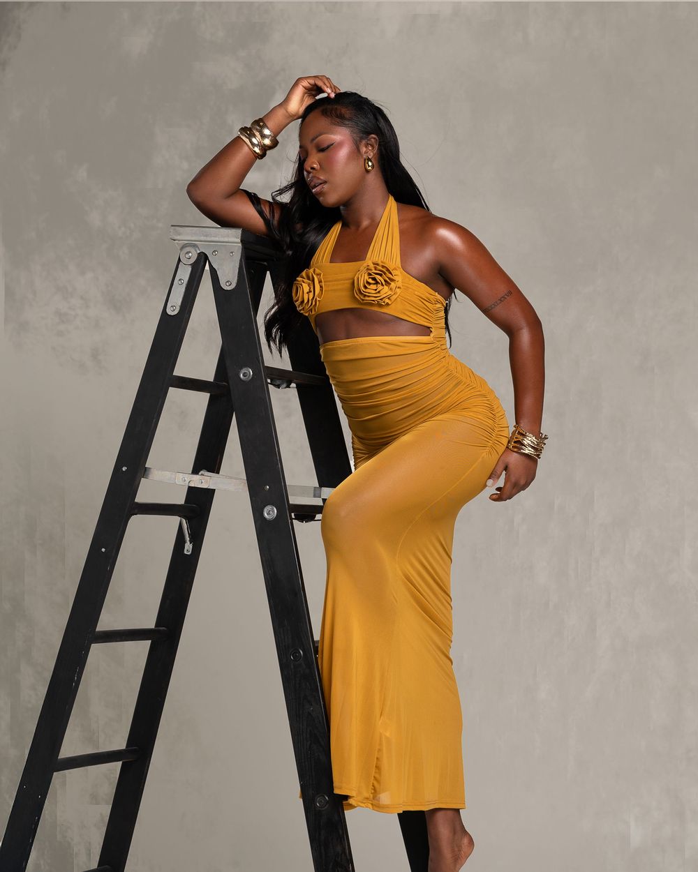 Woman in mustard yellow halter crop top and maxi skirt posing beside a black ladder with gold jewelry.
