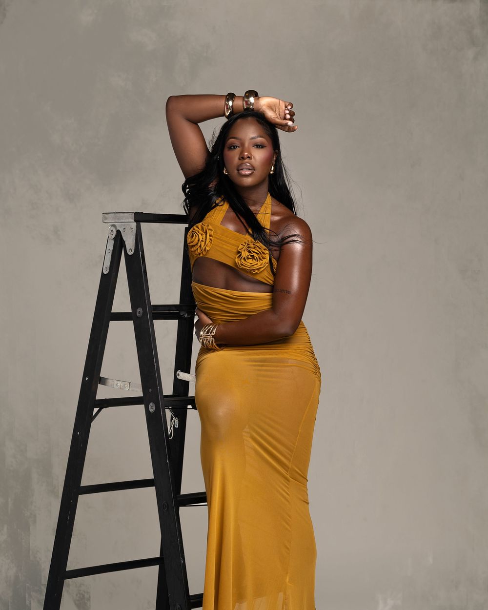 Woman in mustard yellow halter crop top and maxi skirt posing beside a black ladder in a studio.