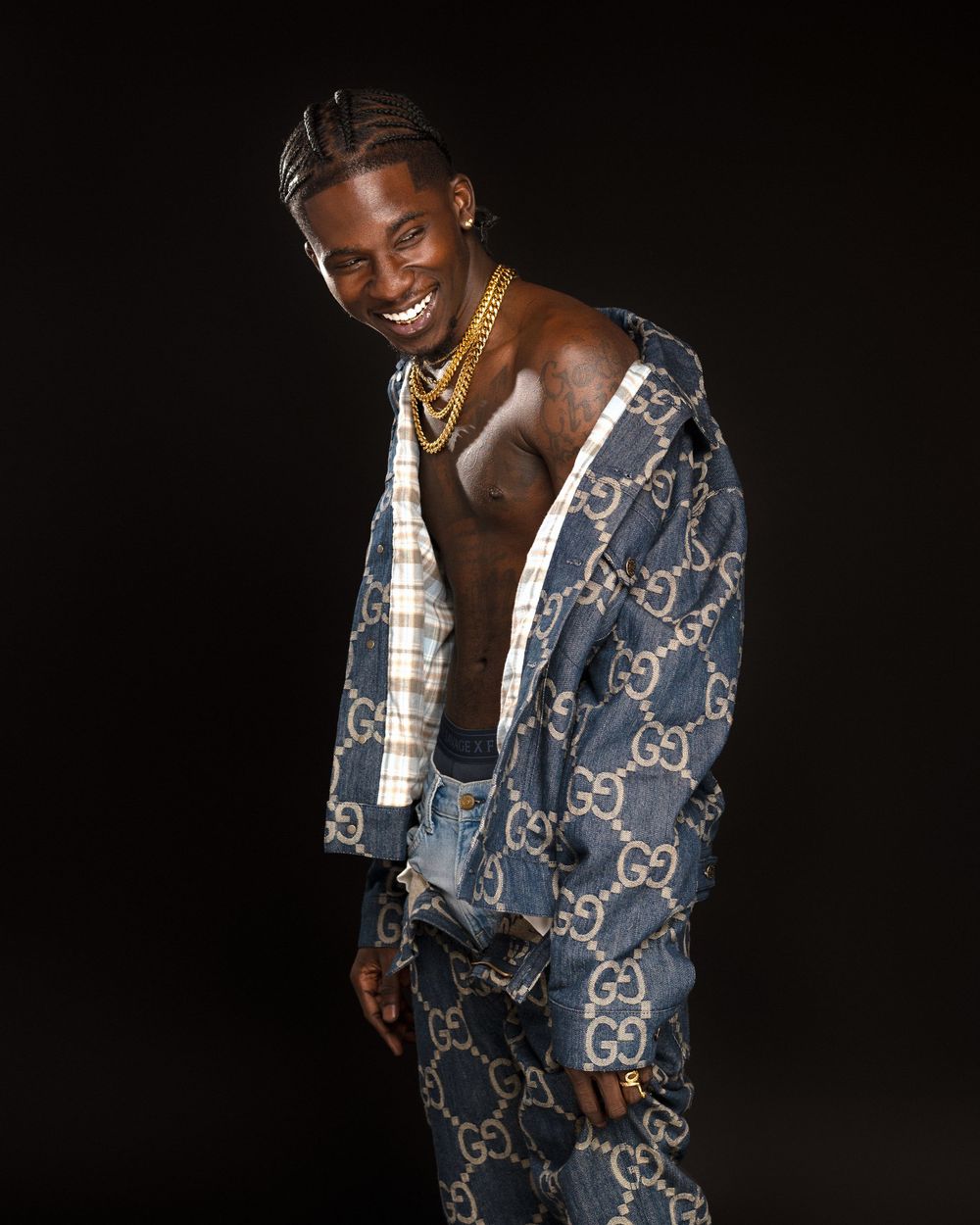 A person wearing a patterned denim jacket with a repeating monogram design poses against a dark background.