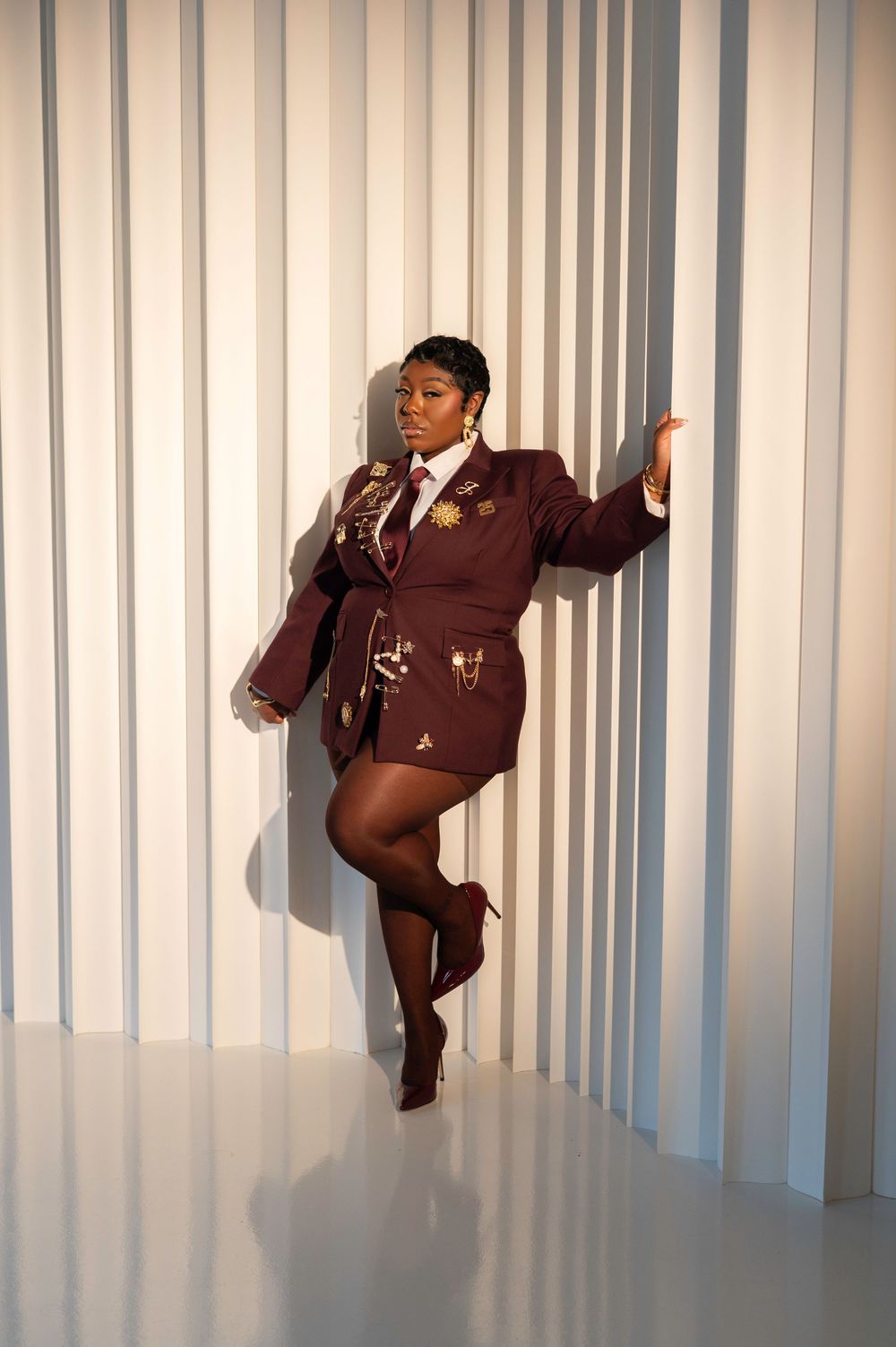 Woman in burgundy blazer dress with gold embellishments, posing confidently against white striped wall in matching heels.