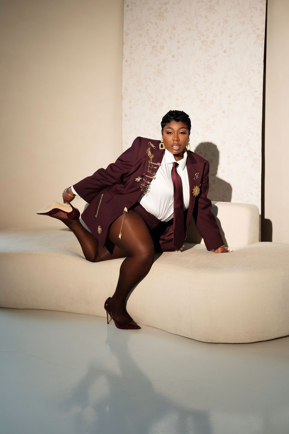 Woman in maroon blazer, white shirt, tie, and heels poses confidently on a cream curved sofa in a bright studio.