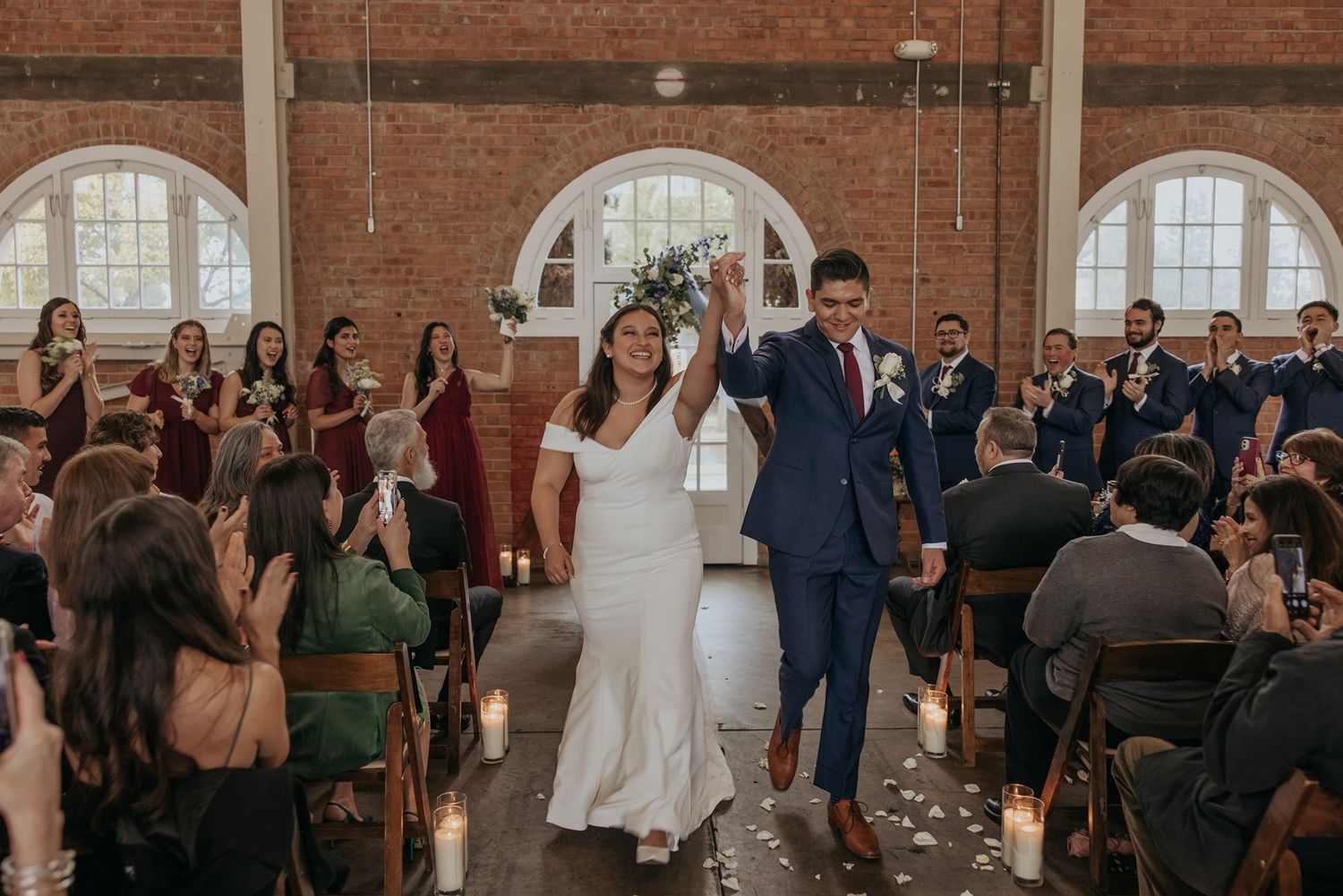 Wedding ceremony in a rustic brick venue with large arched windows and wooden chairs.