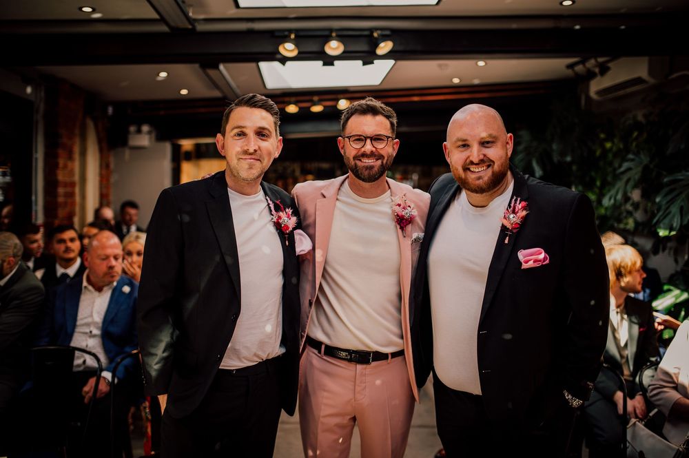 Three men in formal attire smile together at an indoor event with warm lighting and decorative floral boutonnieres.