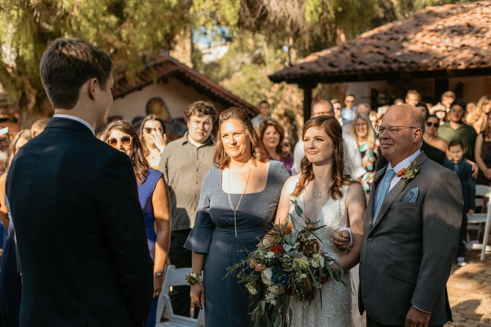 Bride walks down outdoor wedding aisle with bouquet, escorted by family, guests seated in background at Rancho Buena Vista Adobe.