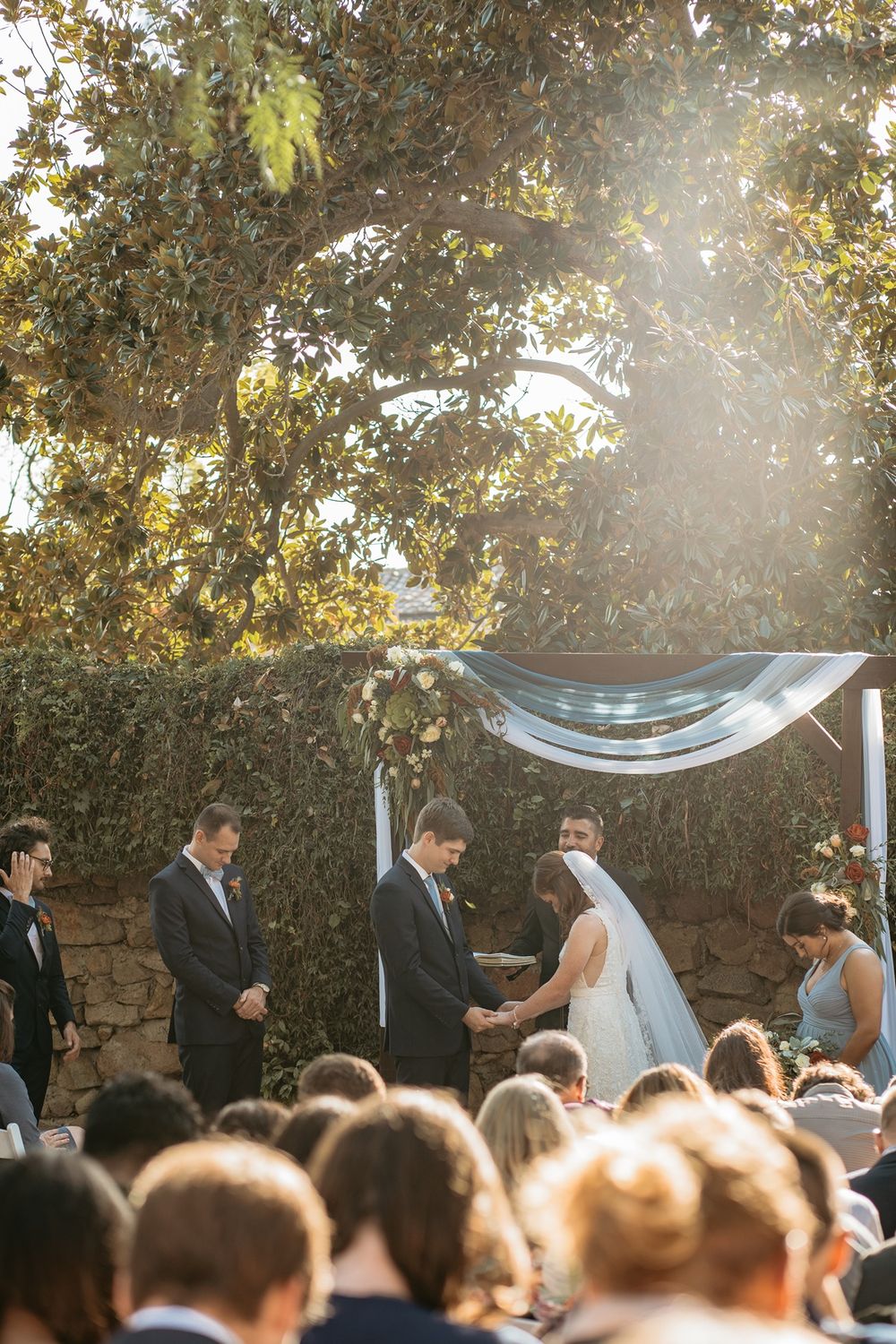 Outdoor wedding ceremony under a sunlit floral arch, bride and groom exchanging vows before seated guests at Rancho Buena Vista Adobe.