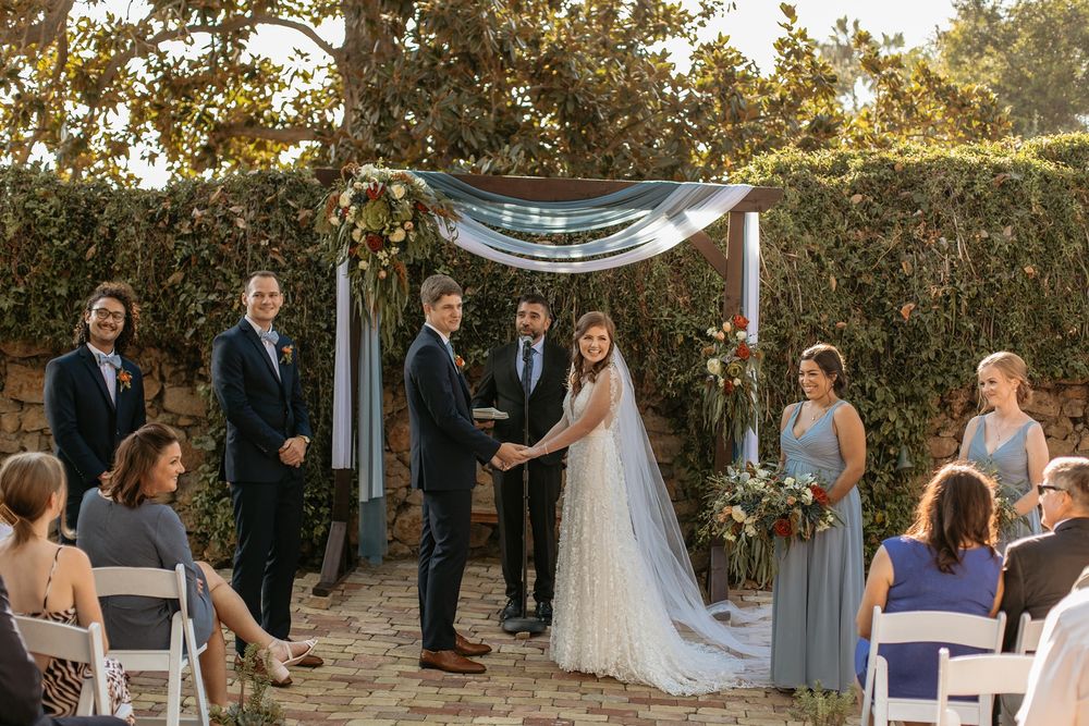 Outdoor wedding ceremony under floral arch with bride and groom holding hands, officiant, and wedding party in blue.