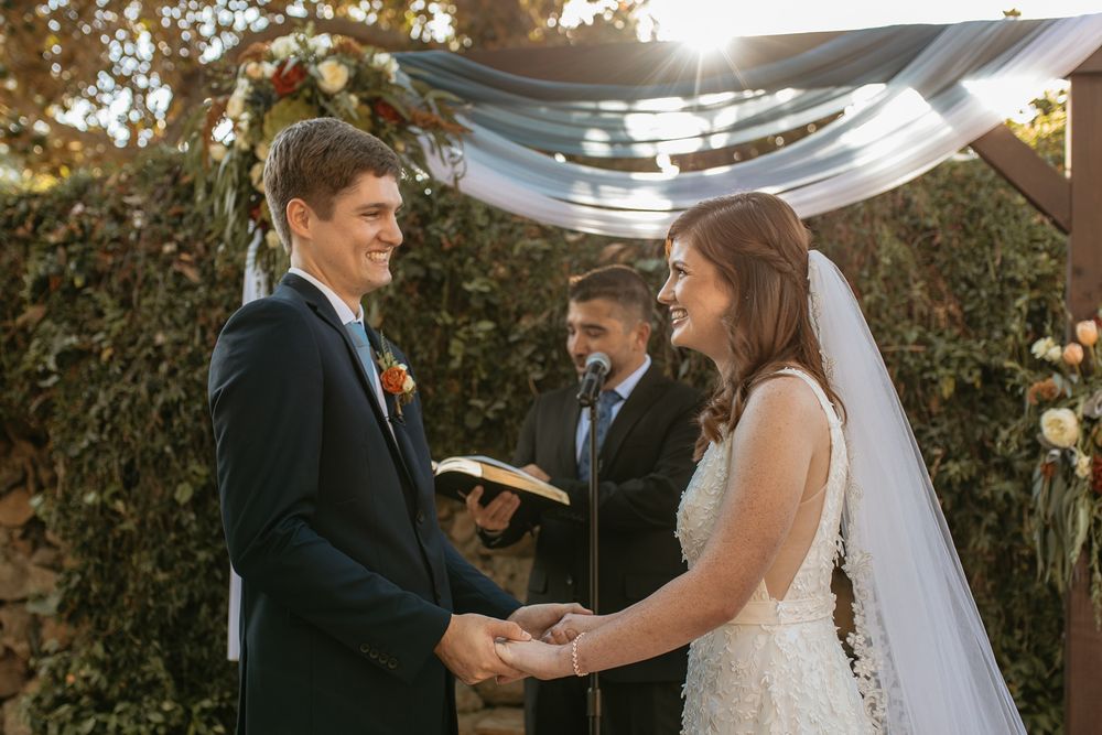 Bride and groom holding hands at outdoor wedding ceremony under floral arch with officiant in background at Rancho Buena Vista Adobe.