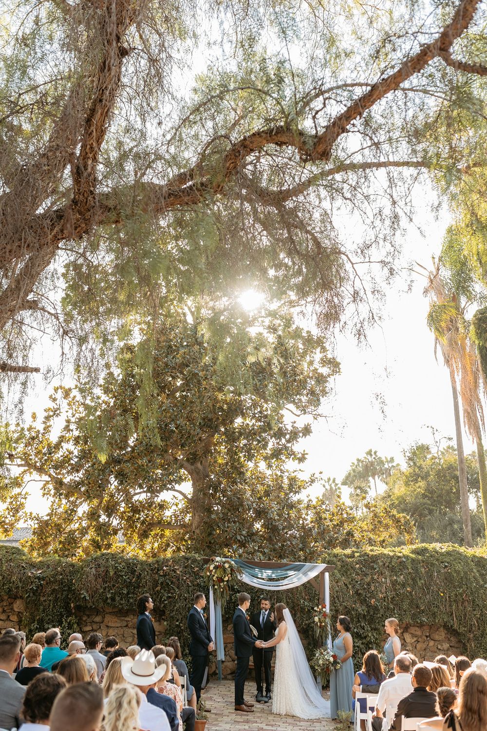 Outdoor wedding ceremony under a large tree with sunlight filtering through, bride and groom at floral arch with seated guests.