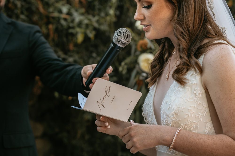 Bride reading wedding vows from elegant card while holding microphone during outdoor ceremony at Rancho Buena Vista Adobe.