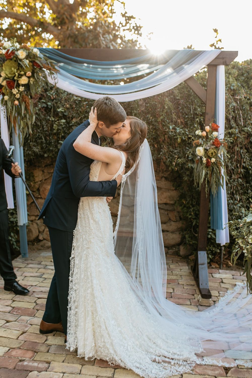 Bride and groom share first kiss under floral arch with blue draping at outdoor wedding ceremony at Rancho Buena Vista Adobe.