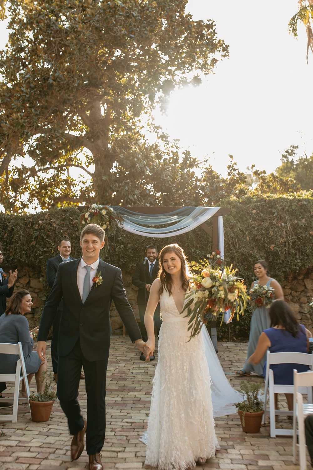 Newlywed couple walks down outdoor wedding aisle, bride in lace gown holding bouquet, groom in dark suit, guests seated nearby.
