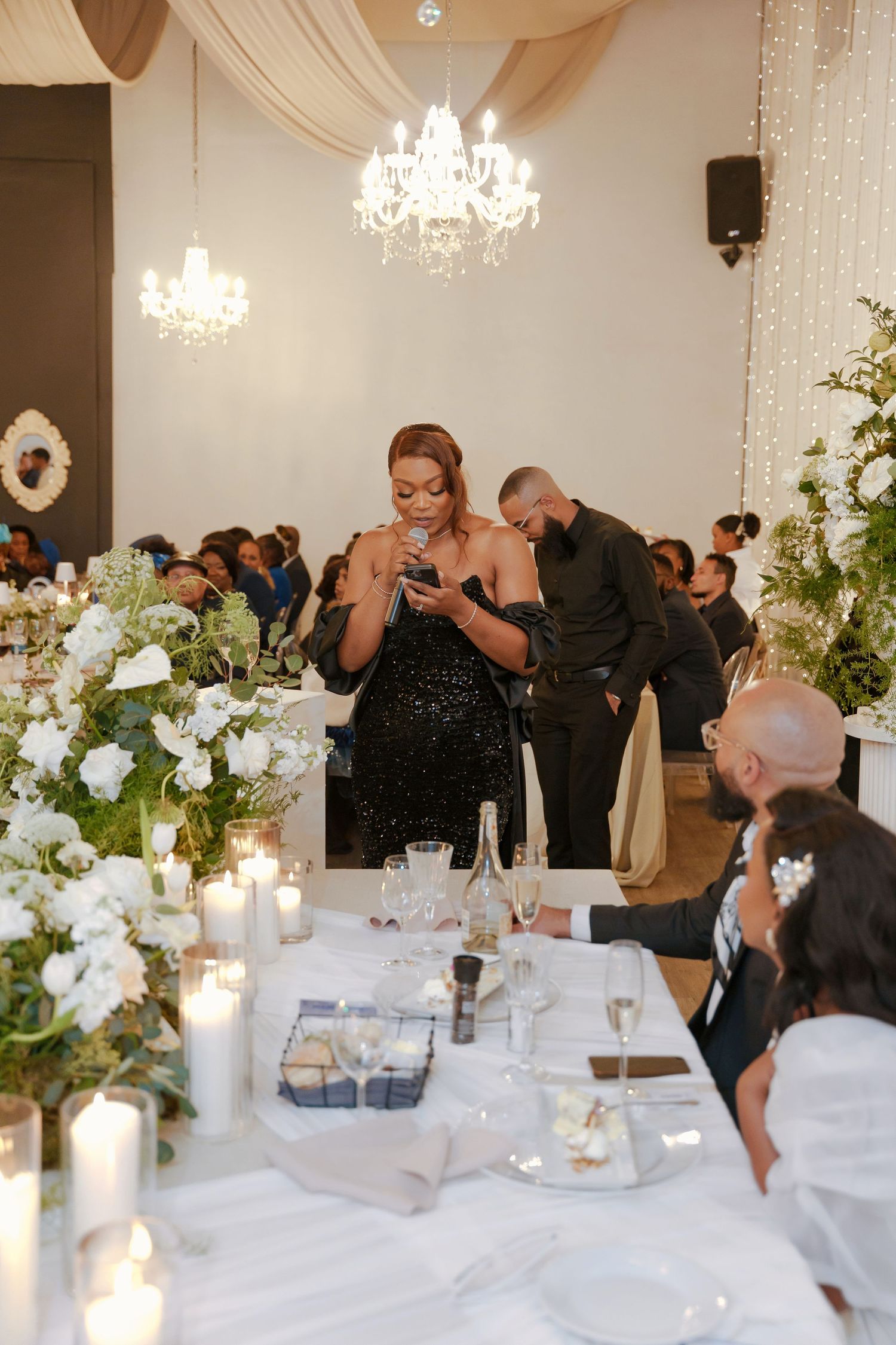 Elegant wedding reception with candlelit tables, white floral arrangements, chandeliers, and guests in formal attire.