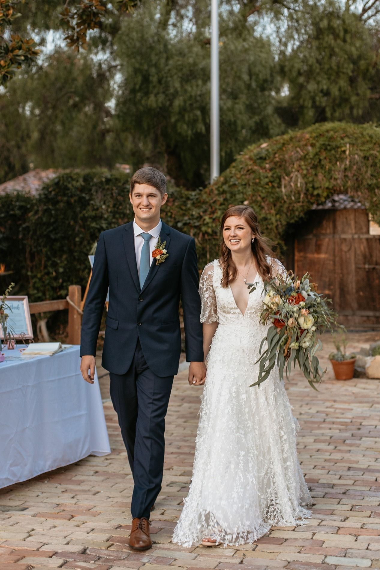 Bride and groom walking hand in hand outdoors, bride in lace gown holding bouquet, groom in navy suit with boutonniere.