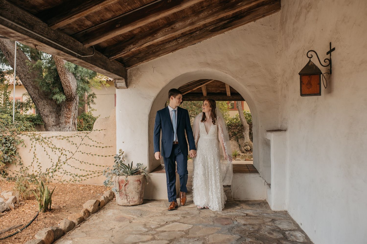 A bride and groom walk hand in hand through a rustic white stucco archway at an outdoor wedding venue.