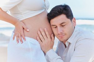 Tender moment as partner listens to pregnant belly on bright beach setting.