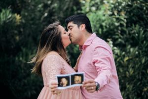 A couple kisses while holding up an ultrasound photo in a garden setting.