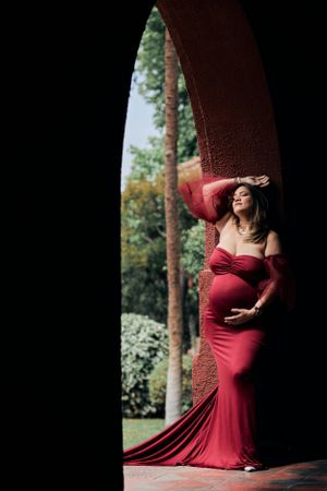 Elegant maternity photo of a woman in flowing red dress posing under a dramatic archway.